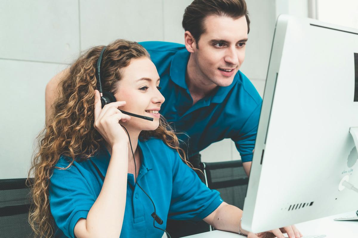 Two support staff at a monitor, one wearing a headset on a call while a colleague looks over her shoulder.
